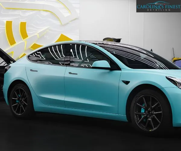 Full side profile of a matte light-blue Tesla Model 3 inside a brightly lit detailing shop