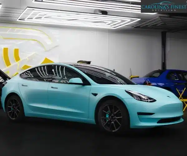 Three-quarter front view of a matte light-blue Tesla Model 3 under overhead garage LED lights.